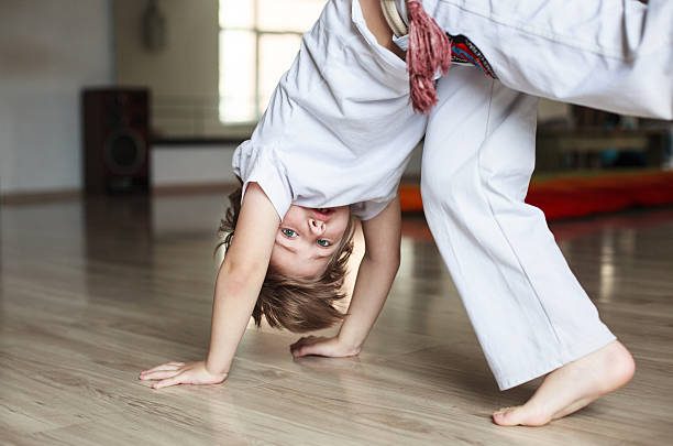 Cute boy in white clothes practicing capoeira (brazilian martial art that combines elements of dance, acrobatics and music)  in gym
