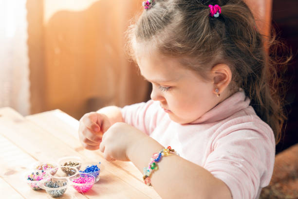 A little girl is engaged in needlework, making jewelry with her own hands, stringing multi-colored beads on a thread.