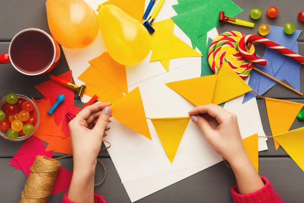 Birthday handmade background. Female hands making party paper decorations and colorful flags garland. DIY accessories on gray wooden planks, top view. Holiday organization, hobb, handicraft concept