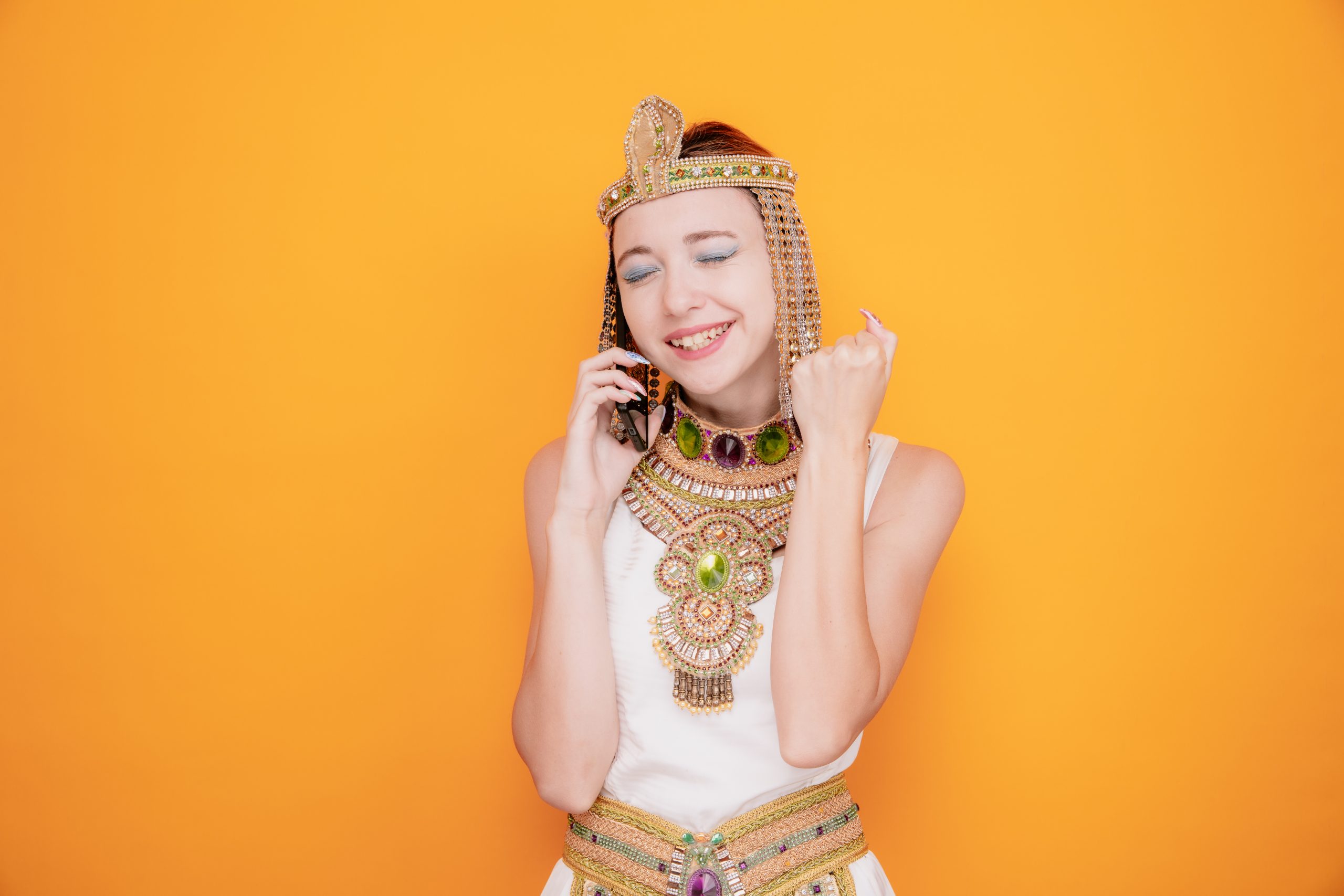 beautiful woman like cleopatra in ancient egyptian costume happy and excited raising fist while talking on mobile phone standing over orange background